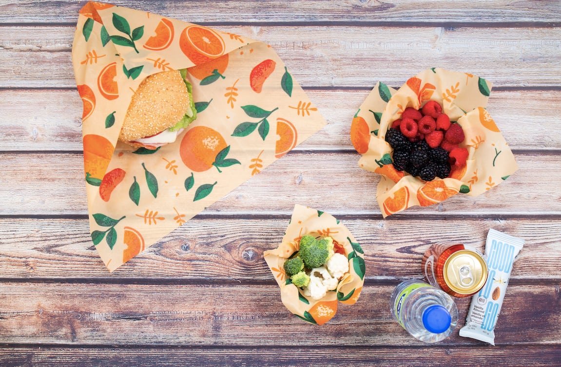 Beeswax food wraps with orange and leaf pattern used to wrap a sandwich, berries, and vegetables on a wooden surface. Drinks and a protein bar are also visible, showcasing a sustainable packed lunch setup.