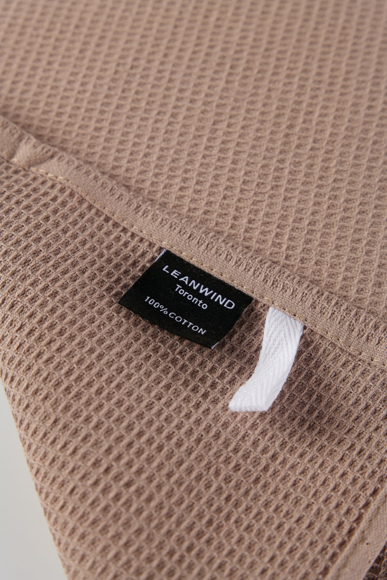 Close-up of a light brown waffle-textured kitchen towel with a black Leanwind Toronto label indicating 100% cotton material. A white hanging loop is visible next to the label.