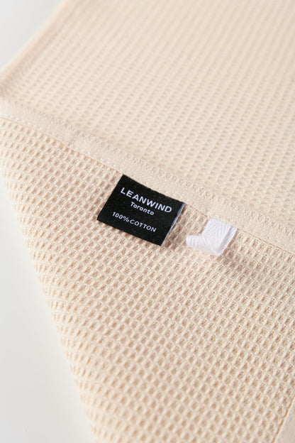 Close-up of a cream-colored waffle-textured kitchen towel with a black label reading &quot;LEANWIND Toronto 100% COTTON&quot; and a white hanging loop attached to the corner.