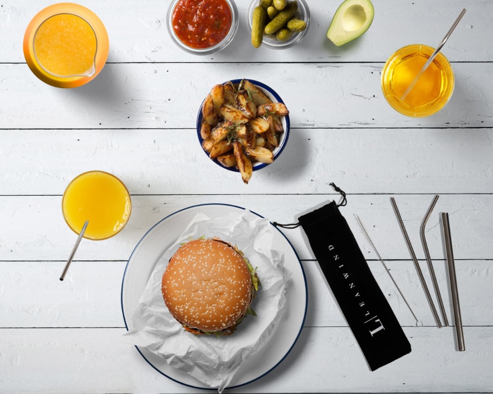 Overhead view of a meal on a white wooden table. The spread includes a hamburger, french fries, orange juice, and various condiments. Leanwind reusable metal straws and a black carrying pouch are visible next to the food items.